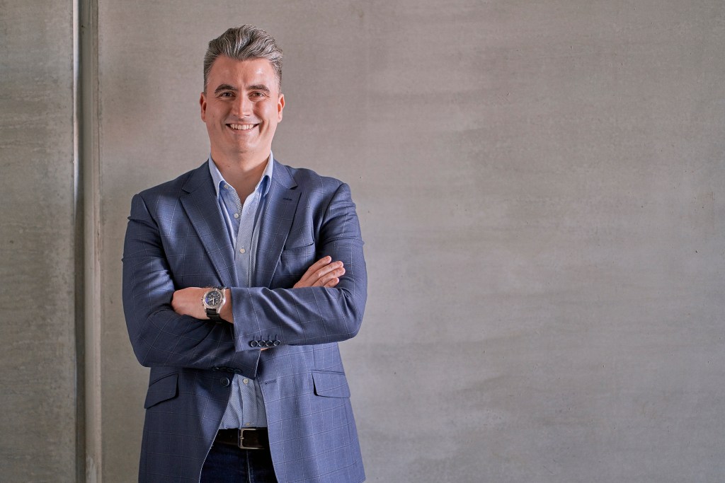 Matt Pearson is standing up and smiling, with his arms folded across his chest. He is wearing a navy pinstripe blazer and a blue shirt underneath.