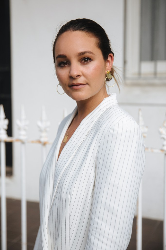 Beck Wadworth, founder of An Organised Life, is pictured wearing a white blazer with black pinstripes. She wears gold earrings and a gold necklace, and is looking at the camera smiling. Her hair is in a bun.