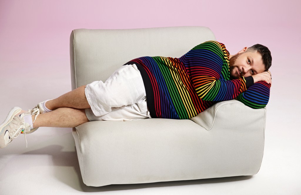 Christian Hull is lying down on a white sofa. He is wearing a rainbow striped jumper and white shorts, and his hand is propped underneath his head.