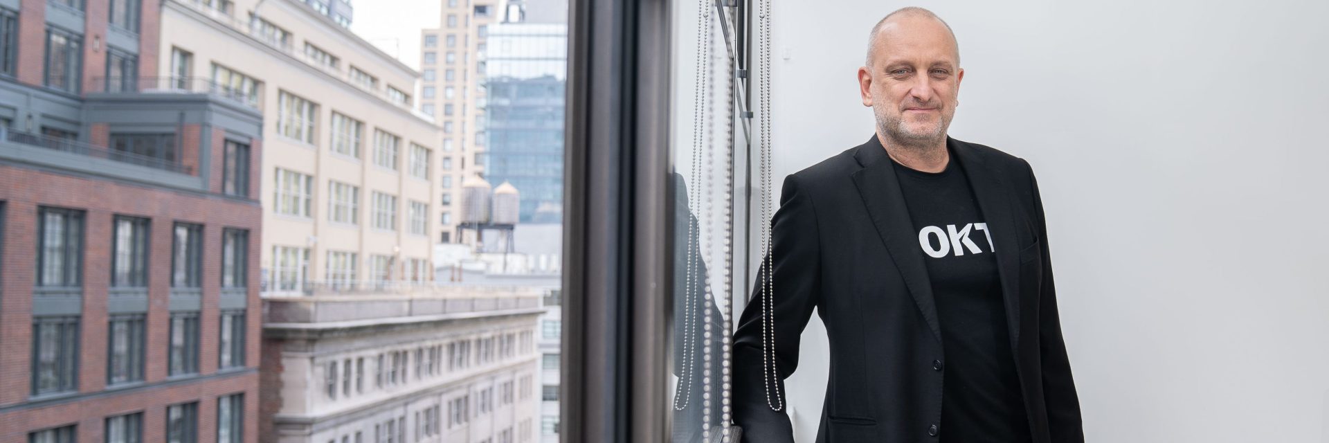 Bruce Buchanan is leaning on a window. He is wearing a black t-shirt that says Rokt underneath a black blazer. Through the window you can see a red brick building in New York.