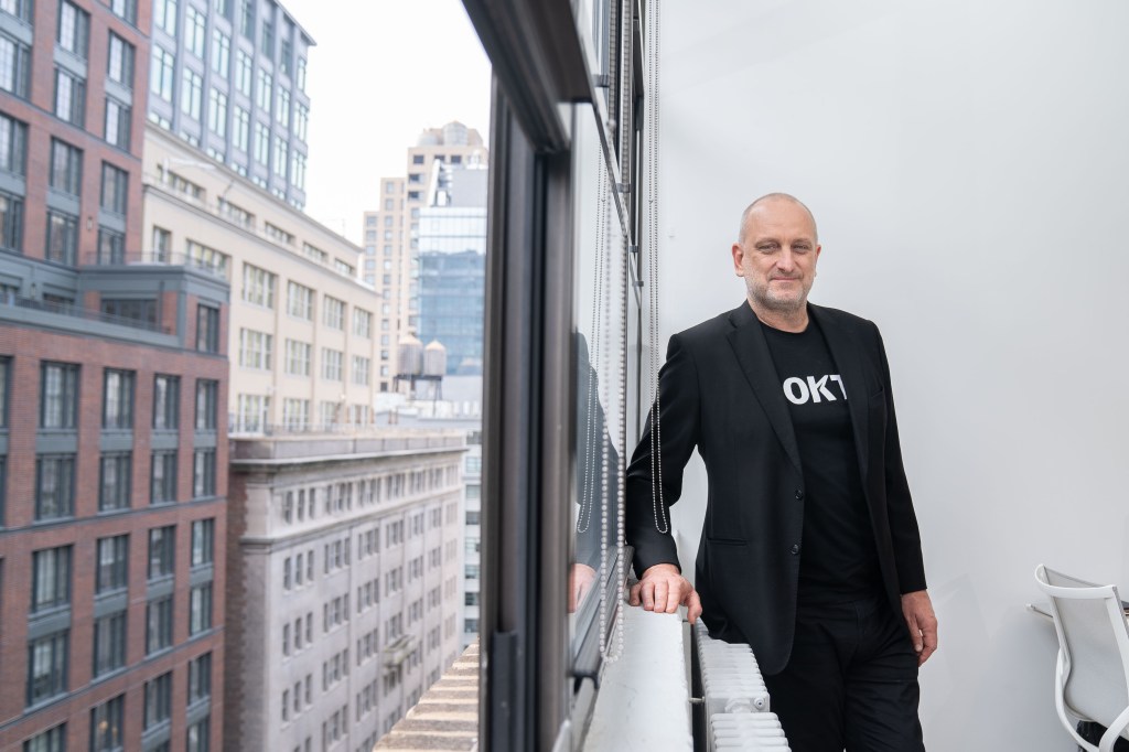 Bruce Buchanan is leaning on a window sill. He is wearing a black t-shirt that says ROKT on it underneath a black blazer. Outside the window, you can see red brick buildings in New York.