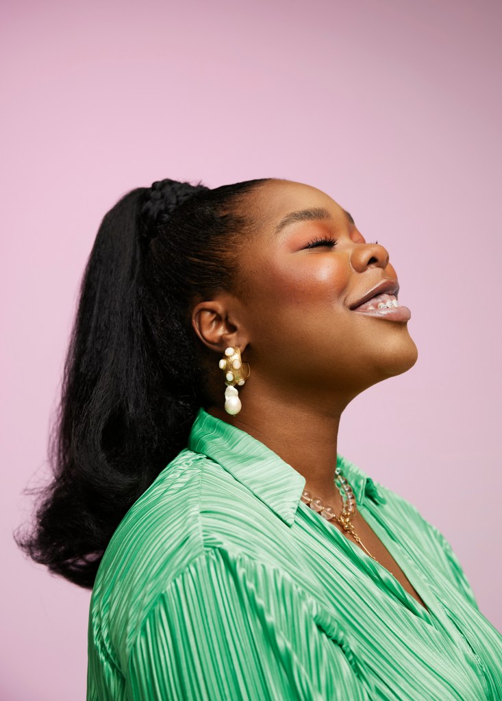 Lilian Ahenkan is pictured wearing a light green plisse top. Her hair is in a ponytail and she is wear gold earrings. She is pictured on her side, and she is smiling up at the sky.