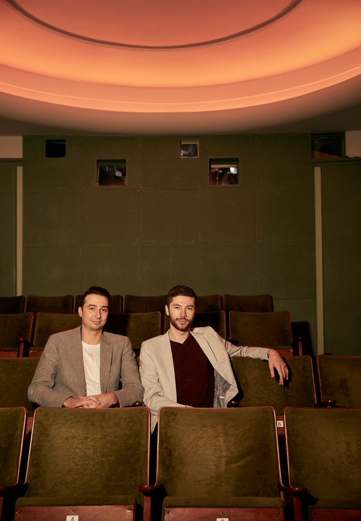 Alex Harper (left) and Angus Goldman (right) are sitting in a movie theatre. They are both wearing blazers with t-shirts underneath. They are looking at the camera.