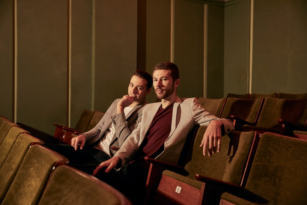 Alex Harper (left) and Angus Goldman (right) are sitting in a movie theatre. They are both wearing blazers with t-shirts underneath. They are looking at the camera.