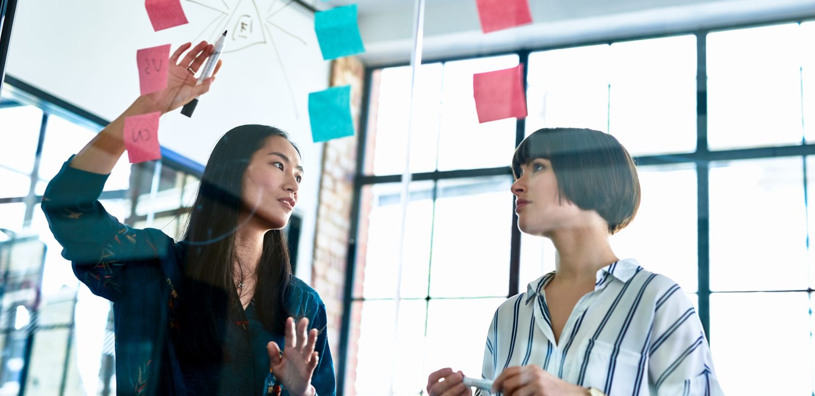 Two women discuss goal setting using sticky notes on a glass wall