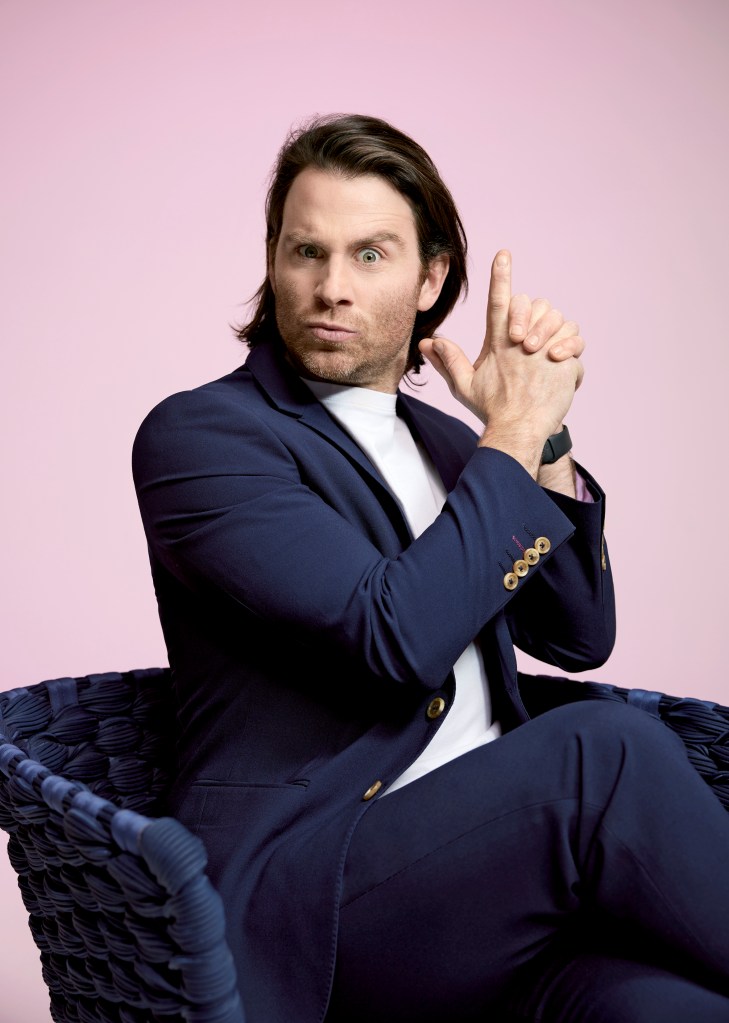 Jimmy Rees is posing on a blue velvet chair. He is wearing a navy suit with a white t-shirt underneath. His hands are making a finger-gun gesture and he is pulling a funny face.