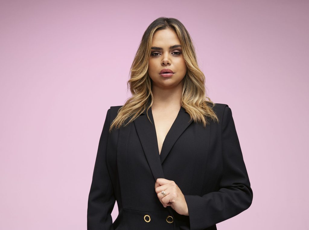 Samantha Harris wears a black suit. She is posing in front of a pink background and her hair is down.