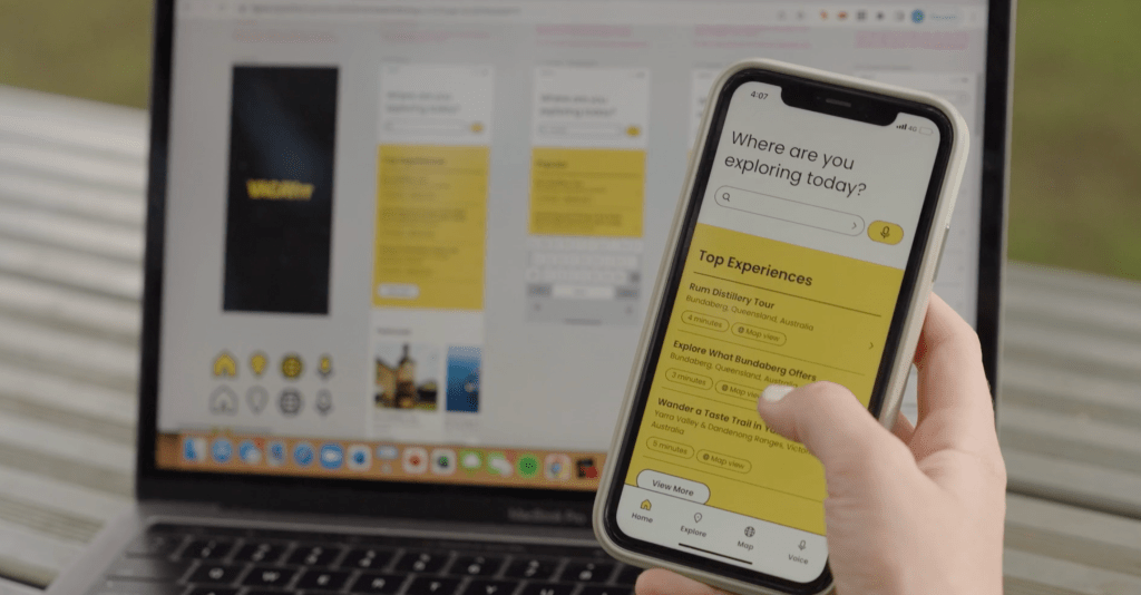 The VacayIT app is open on someone's iPhone. They are holding it, with their finger hovering over a button that says 'Explore what Bundaberg Offers'. In the background is a laptop with the app designs on the screen.