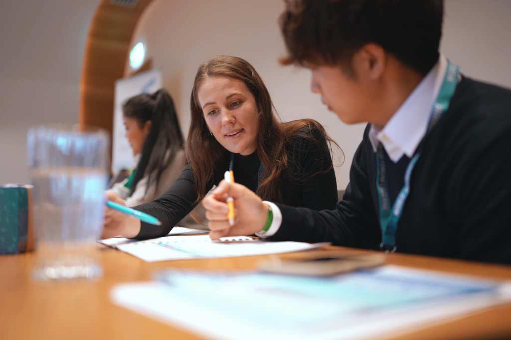 Hailey Brown is pictured at the World Tourism Forum. She is holding a pen, looking down at a piece of paper. She is conversing with someone next to her.