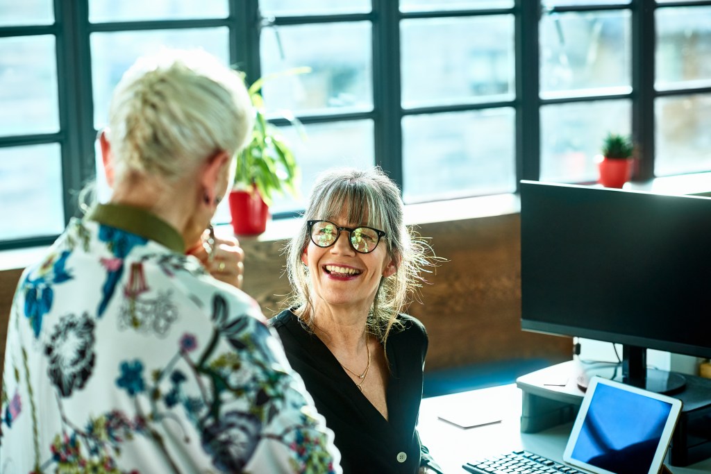 Two women having a conversation at work, woman in her 50s wearing glasses smiling and facing female manager