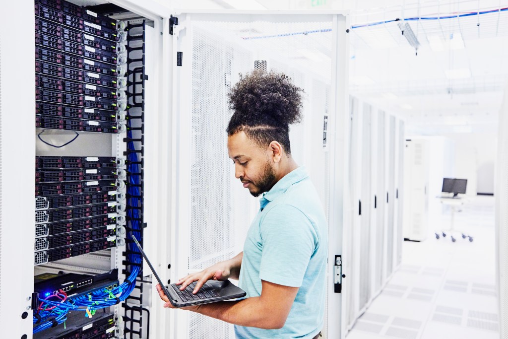 Male IT professional looking at laptop while working on server in data centre