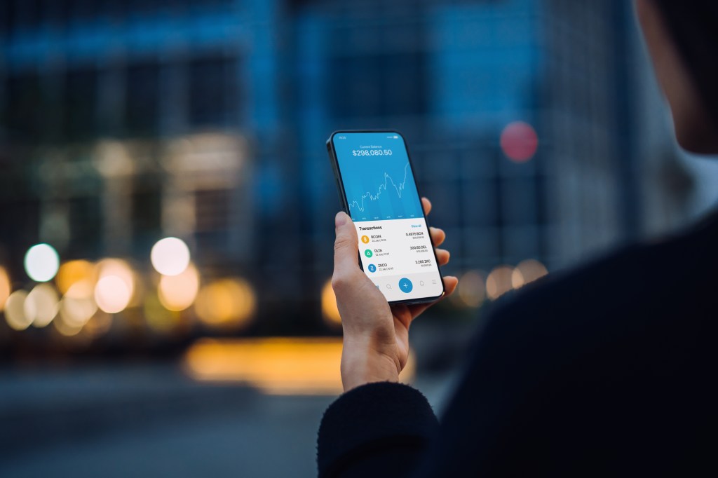 Over the shoulder view of Asian woman using NFT investment wallet on smartphone in city street, working with blockchain technologies, investing or trading NFT (Non-Fungible Token) on cryptocurrency, digital asset, art work and digital ledger