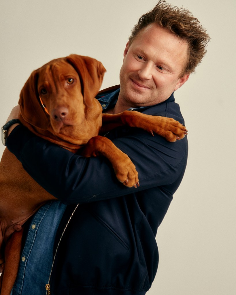 Benjamin Young CEO of Frank Green is holding a dog in his arms. He is wearing a denim shirt with a blue jacket on top. He is smiling.